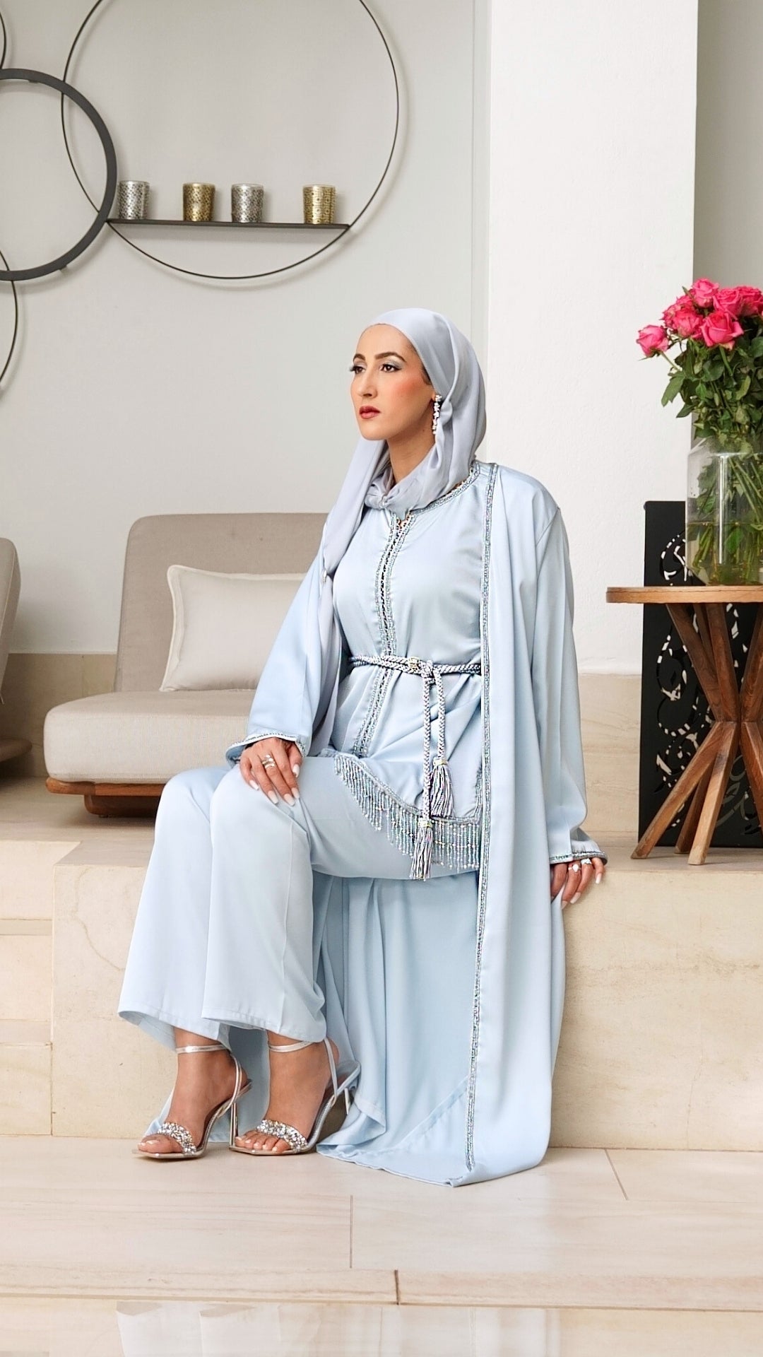 Femme assise portant l'ensemble Jabador Rihem bleu ciel. Tenue 3 pièces moderne avec kimono, tunique et pantalon, ornée de franges et finitions argentées. Création Beldyness