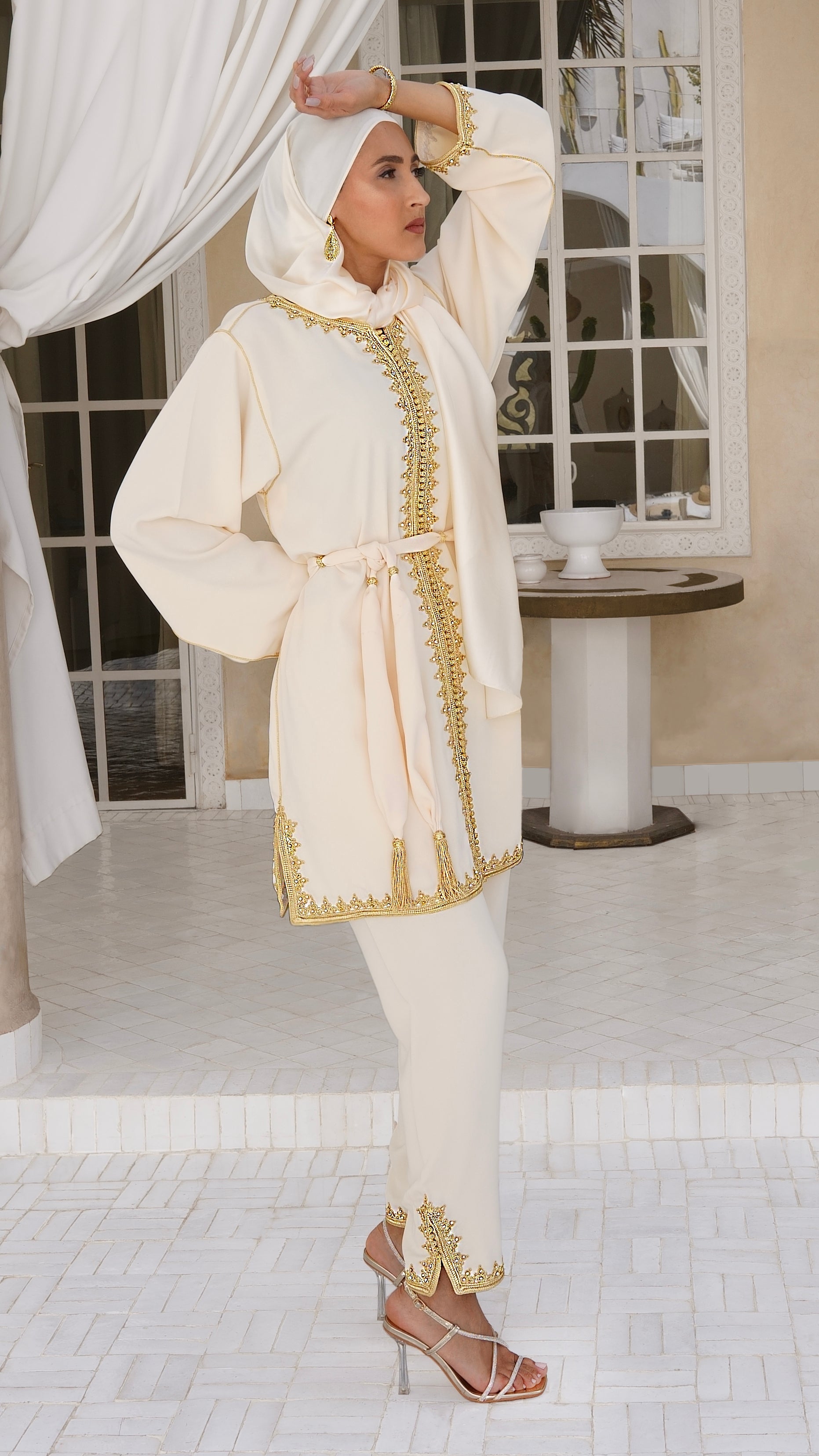 Mannequin posant debout avec le Jabador johar beige crème. Un ensemble marocain 2 pièces composé d'une tunique croisée avec ceinture à pompons et d'un pantalon fluide. Finitions artisanales en broderie dorée et perles pour une allure chic et pudique signée Beldyness.