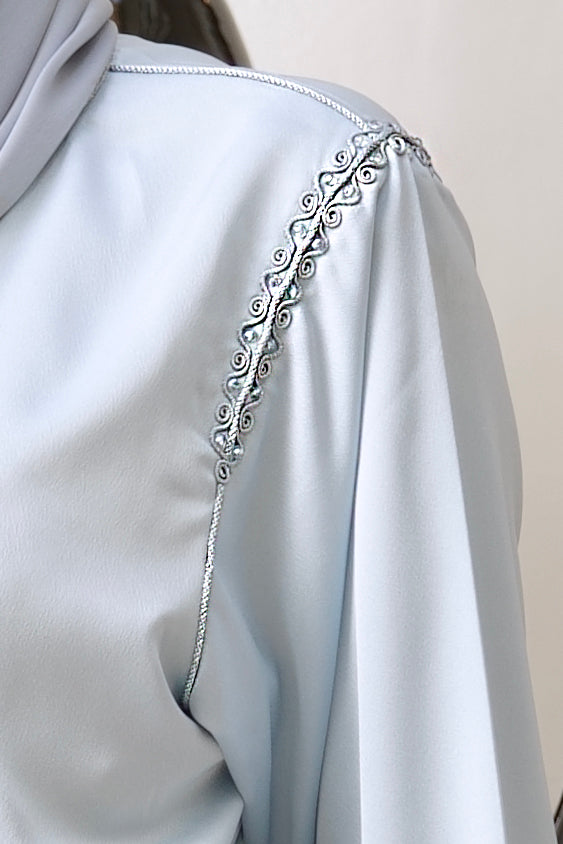 Zoom sur la broderie argentée à l'épaule du Jabador Rihem. Détail des finitions minutieuses et des incrustations de cristaux sur tissu bleu ciel.