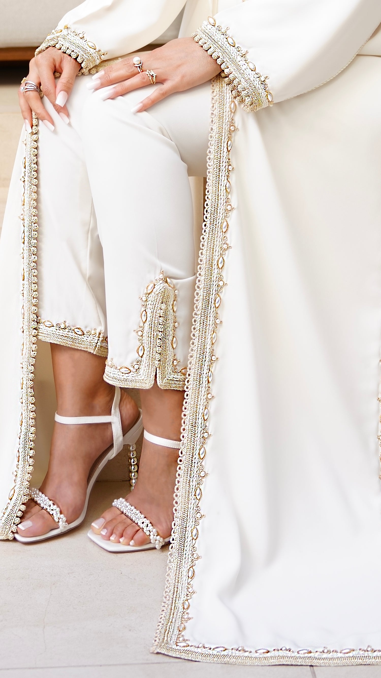 Gros plan sur les finitions d'exception de l'ensemble caftan-pantalon Azhar Ivoire. Détail de la fente au niveau des chevilles, sublimée par des broderies dorées et des incrustations de perles nacrées. L'alliance parfaite du confort et de l'élégance pour une mariée, signée Beldyness.